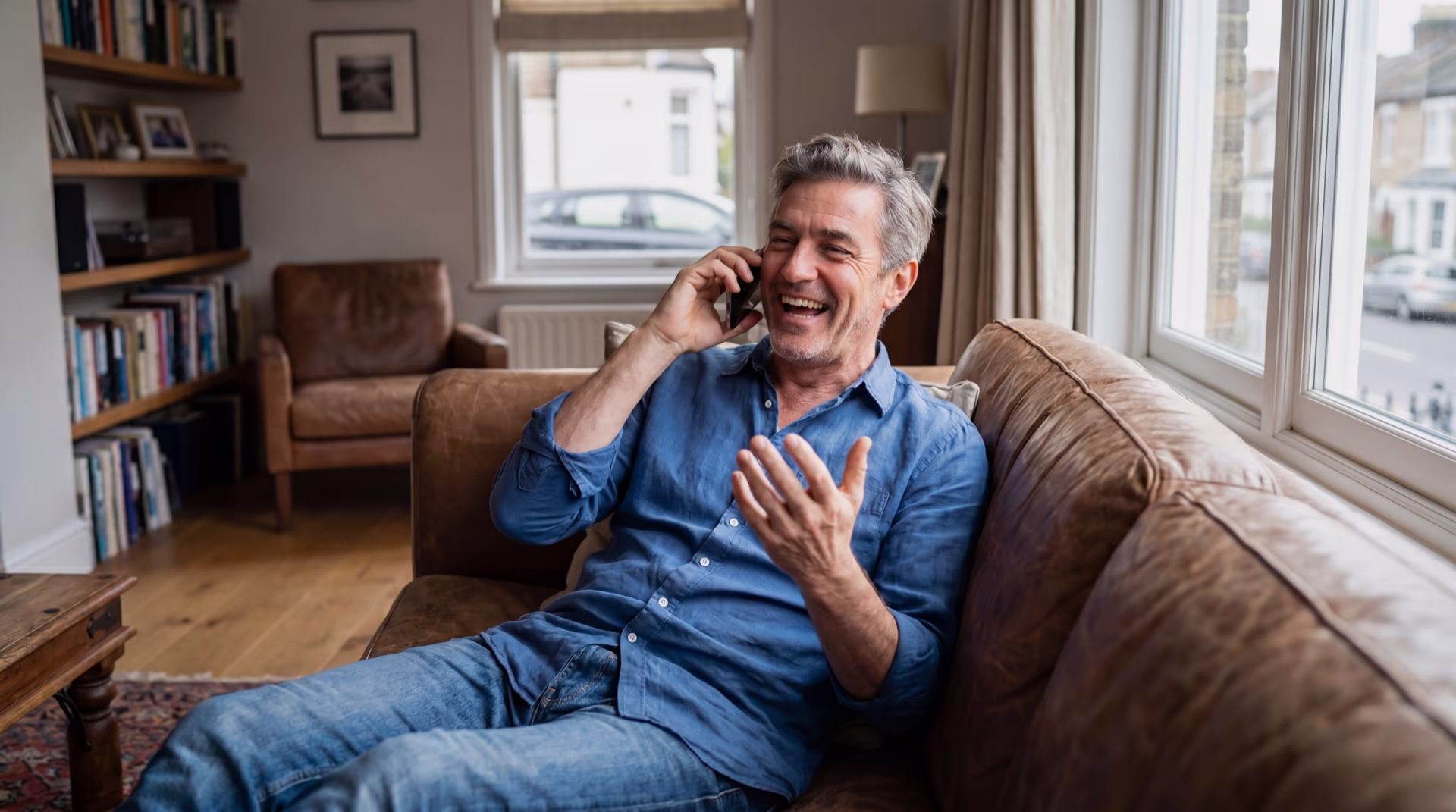 A grey-haired man in a denim shirt laughs while talking on the phone at home